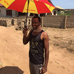 Ghanaian journalist Latif Iddrisu stands outside his Accra home in May 2018, wearing a neck brace and carrying an umbrella because direct sunlight aggravated his head injury from an attack by police in March 2018. (CPJ/Jonathan Rozen)