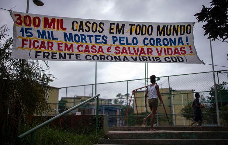 Um banner dizendo “300milasos em todo mundo, 15 mil mortes pelo corona! Ficar em casa é salvar vidas!” pendurado na Cidade de Deus, favela do Rio de Janeiro, em 7 de abril de2020, durante a pandemia de COVID-19. Os jornalistas comunitários doRioenfrentan desafios diários para informar os moradores sobre a COVID-19. (AFP/Mauro Pimentel)