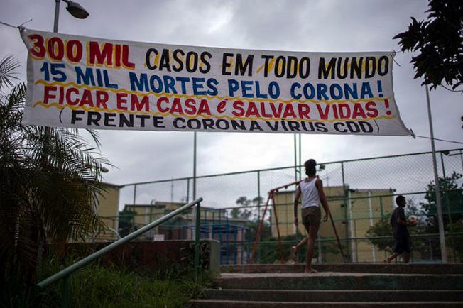 Um banner dizendo “300milasos em todo mundo, 15 mil mortes pelo corona! Ficar em casa é salvar vidas!” pendurado na Cidade de Deus, favela do Rio de Janeiro, em 7 de abril de2020, durante a pandemia de COVID-19. Os jornalistas comunitários doRioenfrentan desafios diários para informar os moradores sobre a COVID-19. (AFP/Mauro Pimentel)