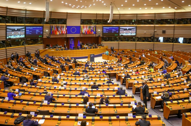 Lawmakers are shown seated at desks in rows facing a podium and EU flags in a large parliamentary building.