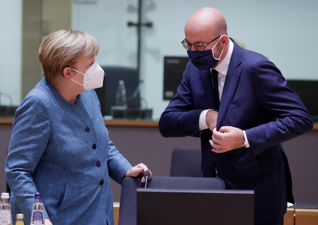 Two politicians wearing masks are seen in conversation behind a desk.