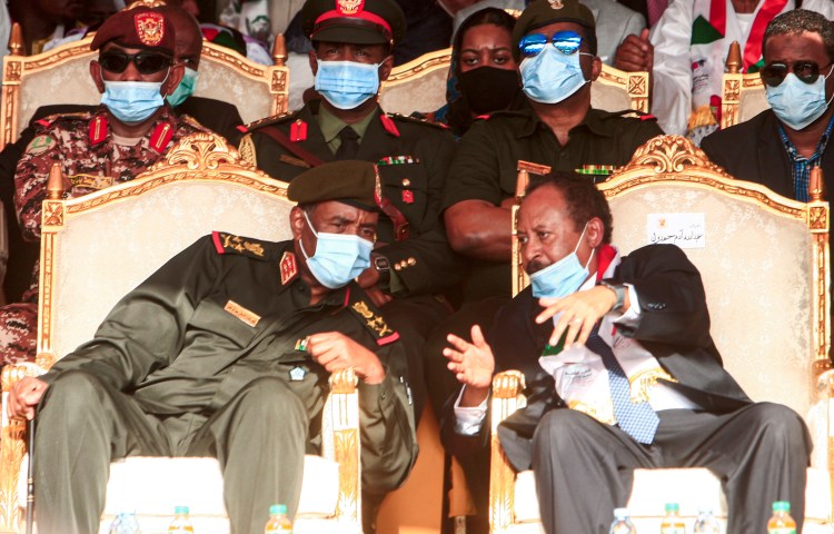 A man in military uniform and a coronavirus mask seated in an elaborate chair leans over to talk with a man in a suit and a coronavirus mask in the neighboring chair.