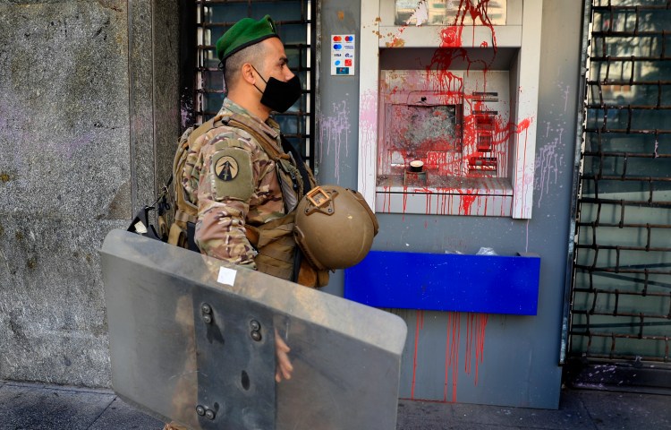 A soldier wearing a mask is shown in profile passing an ATM machine covered in red paint.