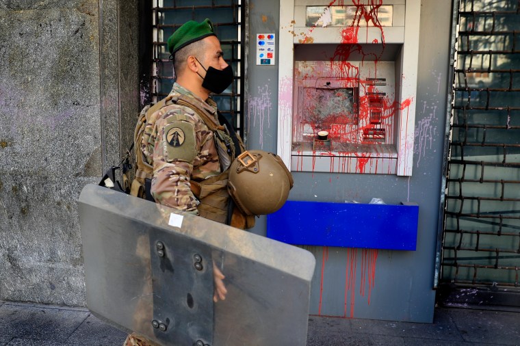 A soldier wearing a mask is shown in profile passing an ATM machine covered in red paint.