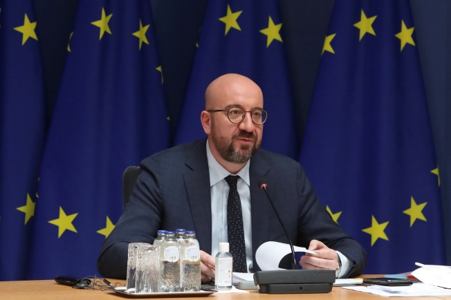 A seated man in a suit speaks into a microphone against a backdrop of the EU flag.