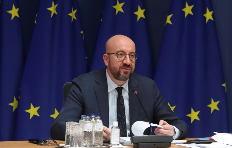 A seated man in a suit speaks into a microphone against a backdrop of the EU flag.