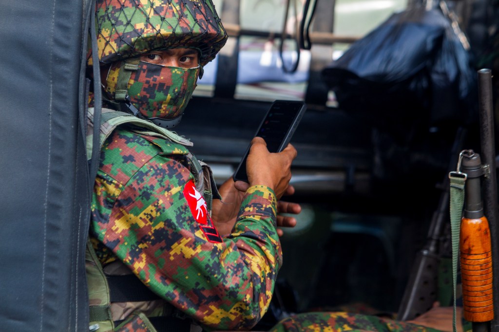 A man in military fatigues and a face mask holding a cell phone in his hand looks at the camera.