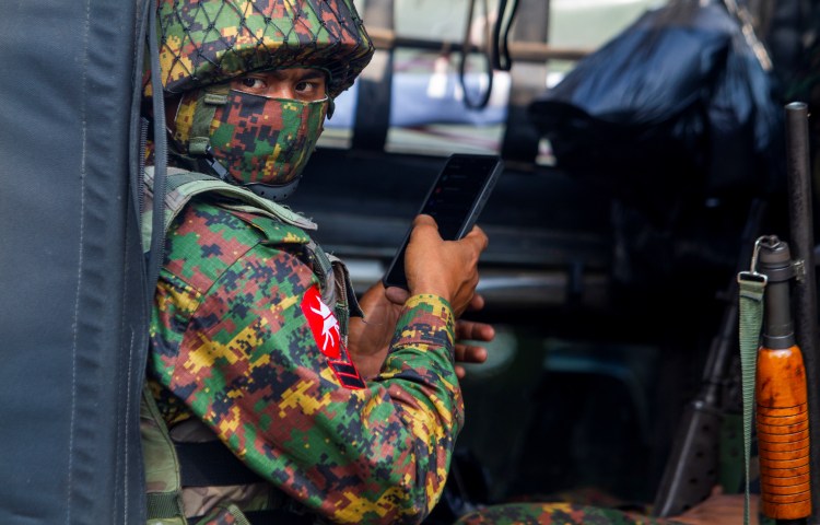 A man in military fatigues and a face mask holding a cell phone in his hand looks at the camera.