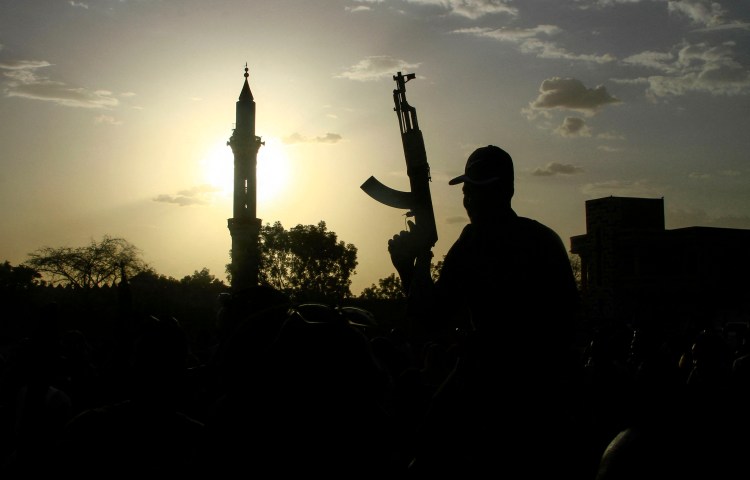 A fighter holds up a gun backdropped by the minaret of a mosque and the sun.