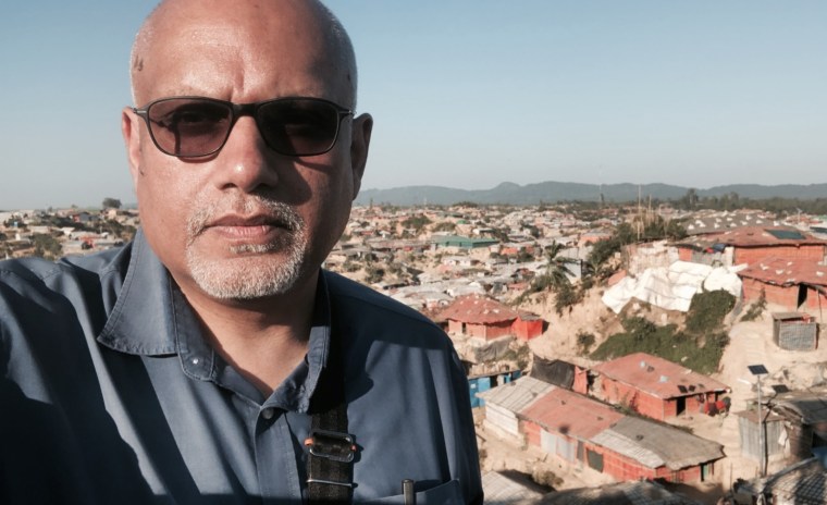 A man in a blue shirt takes a selfie in front of houses.