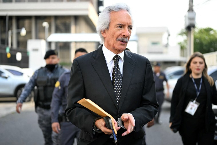 A handcuffed man in a suit walks carrying folders.