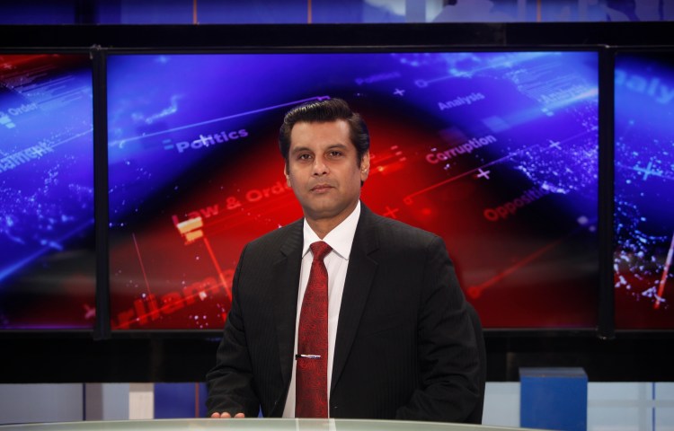 A man in a suit stands before a news broadcast background.