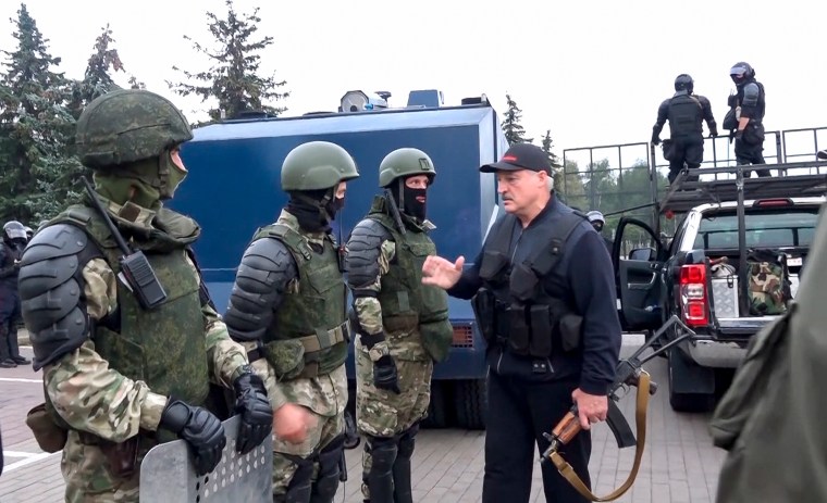 A man carrying a rifle talks to three men in green military gear.