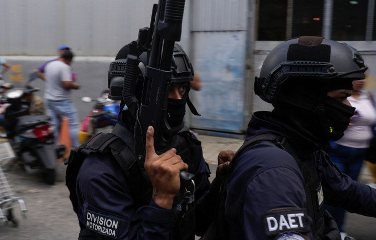 Two officers in riot gear ride past people on a street on a motorcycle. One is carrying a rifle.