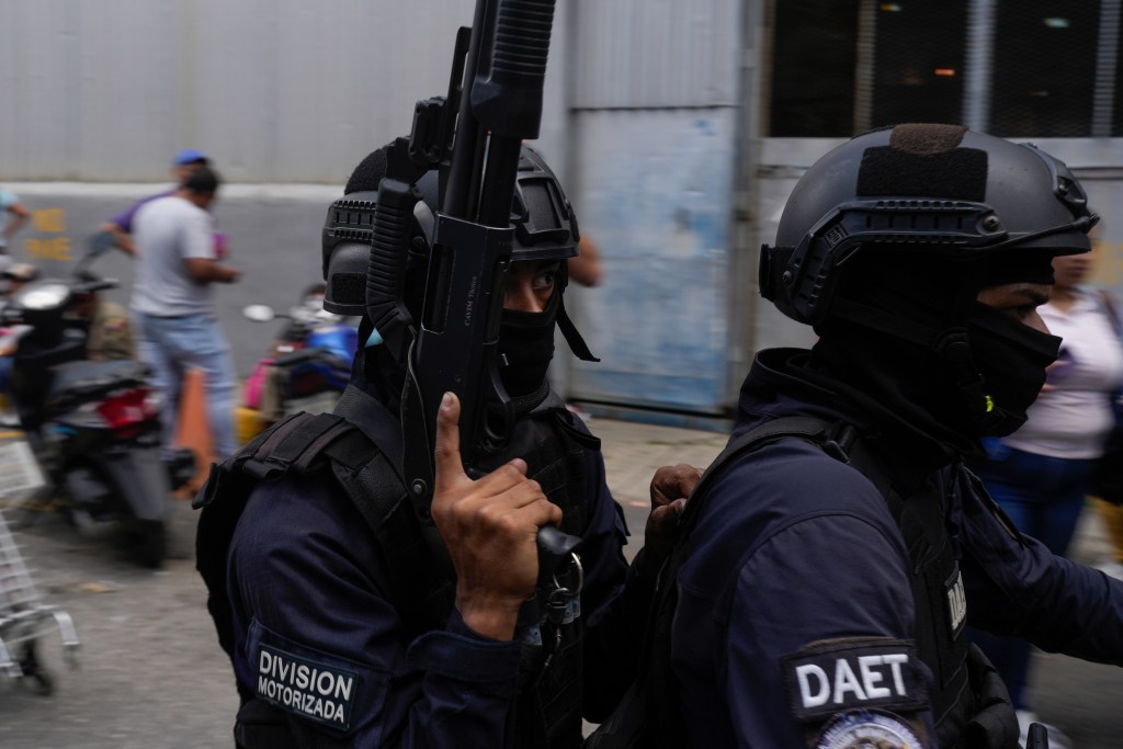 Two officers in riot gear ride past people on a street on a motorcycle. One is carrying a rifle.