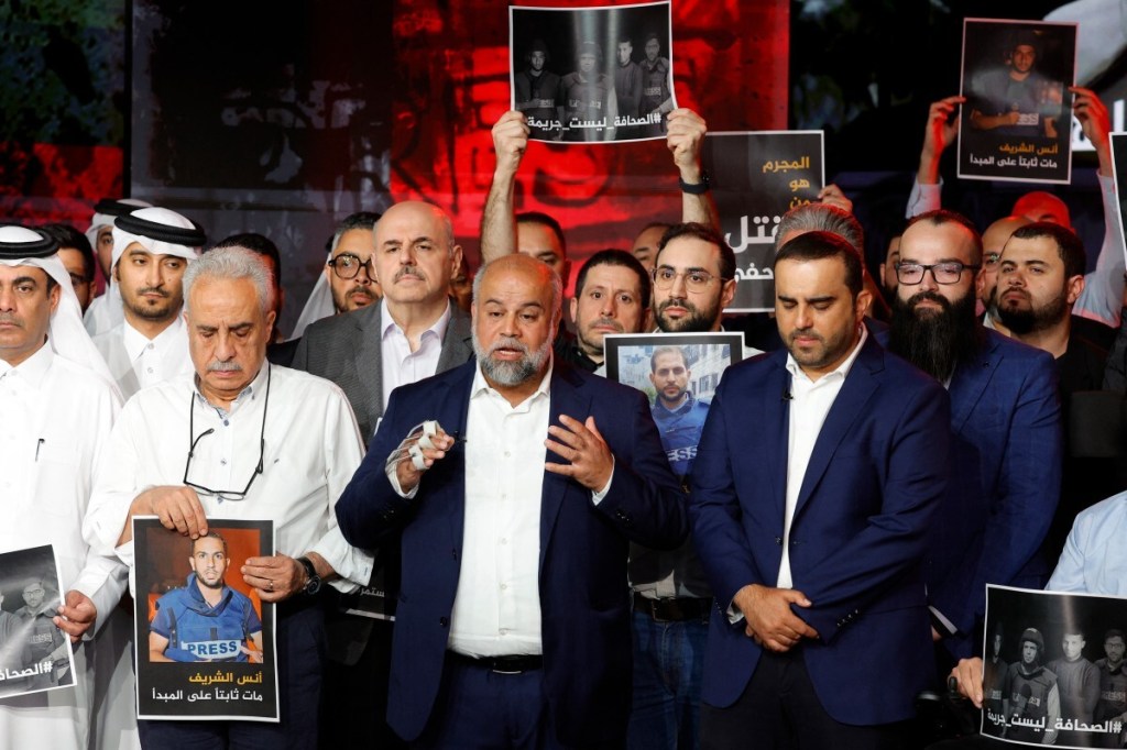 A somber group of men in formal dress stand together facing the camera, some holding posters and images of Al Jazeera staff recently killed in an Israeli strike. 